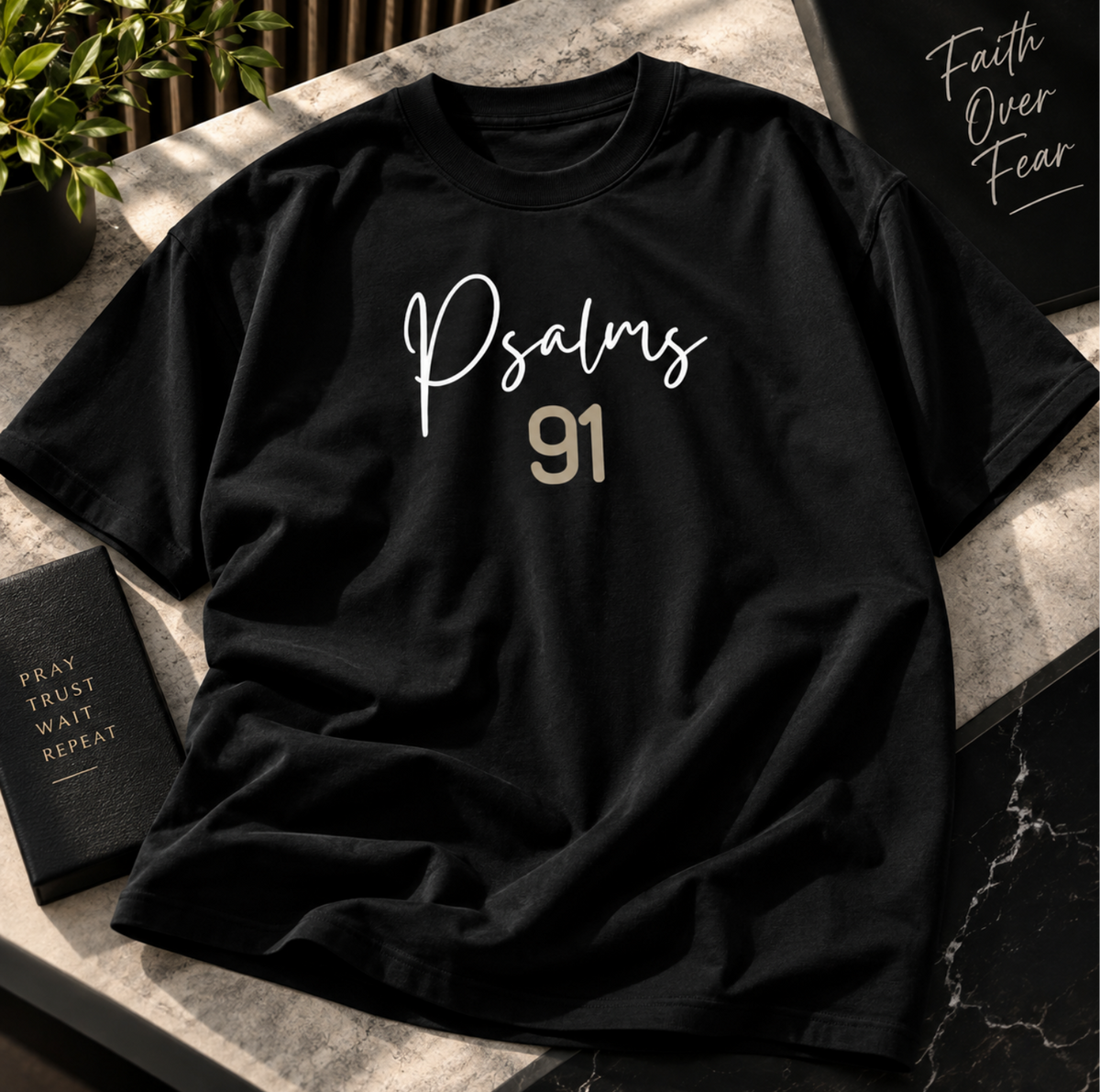 A black short-sleeve T-shirt laid flat on a stone surface, featuring the word 'Psalms' in white script and the number '91' in a metallic gold tone across the chest. Decorative books and plants are partially visible around it.