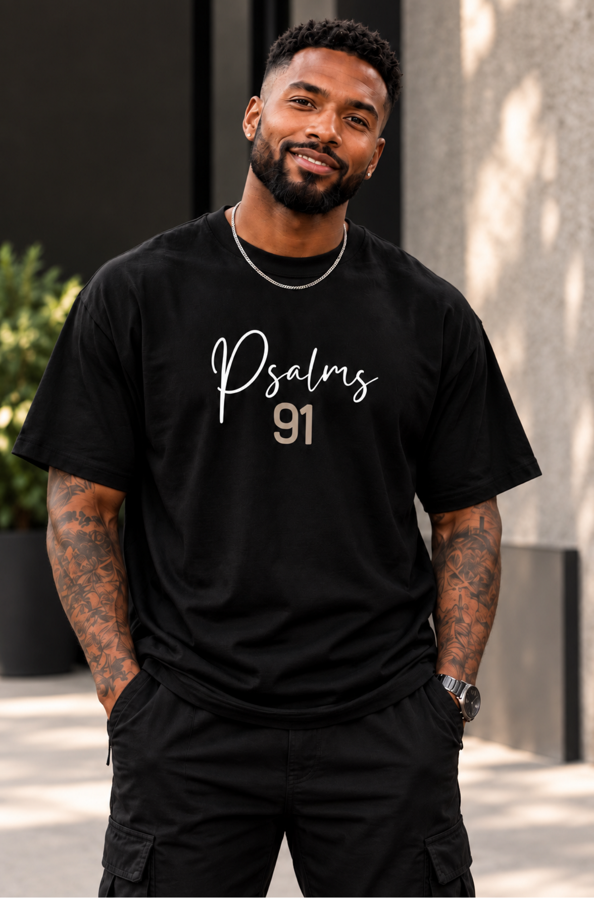 A man wearing a black graphic t-shirt with 'Psalms 91' printed in white and gold lettering, standing outdoors against a modern concrete wall.