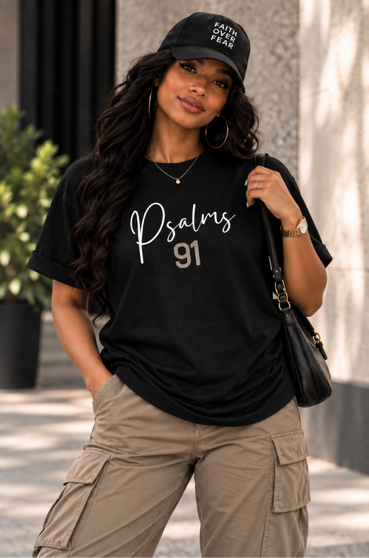 A woman wearing a black graphic t-shirt with 'Psalms 91' printed in white and beige, paired with khaki cargo pants, a black cap, and a black shoulder bag, posing outdoors.