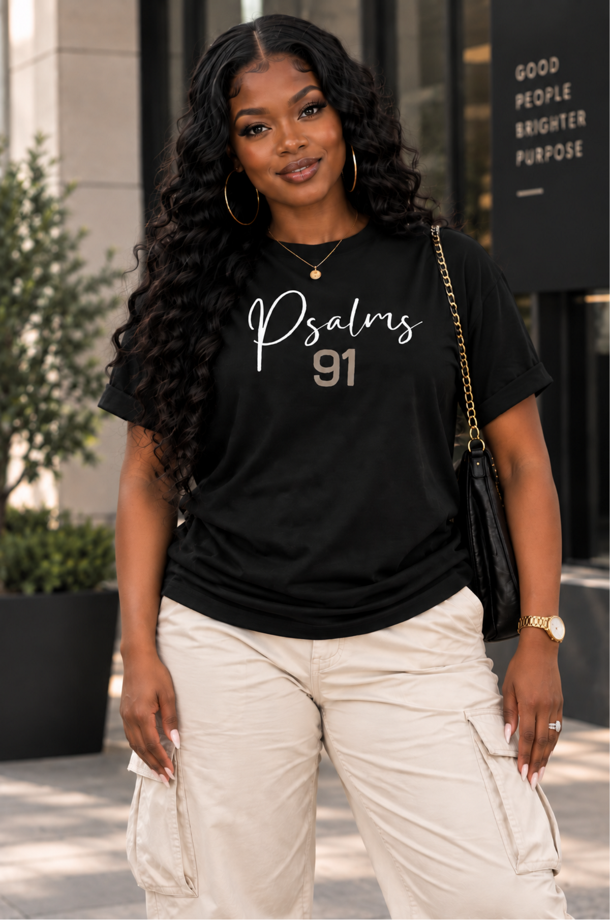 A woman wearing a black graphic t-shirt with 'Psalms 91' printed in white and gold, paired with beige cargo pants, standing outdoors in front of a modern building.