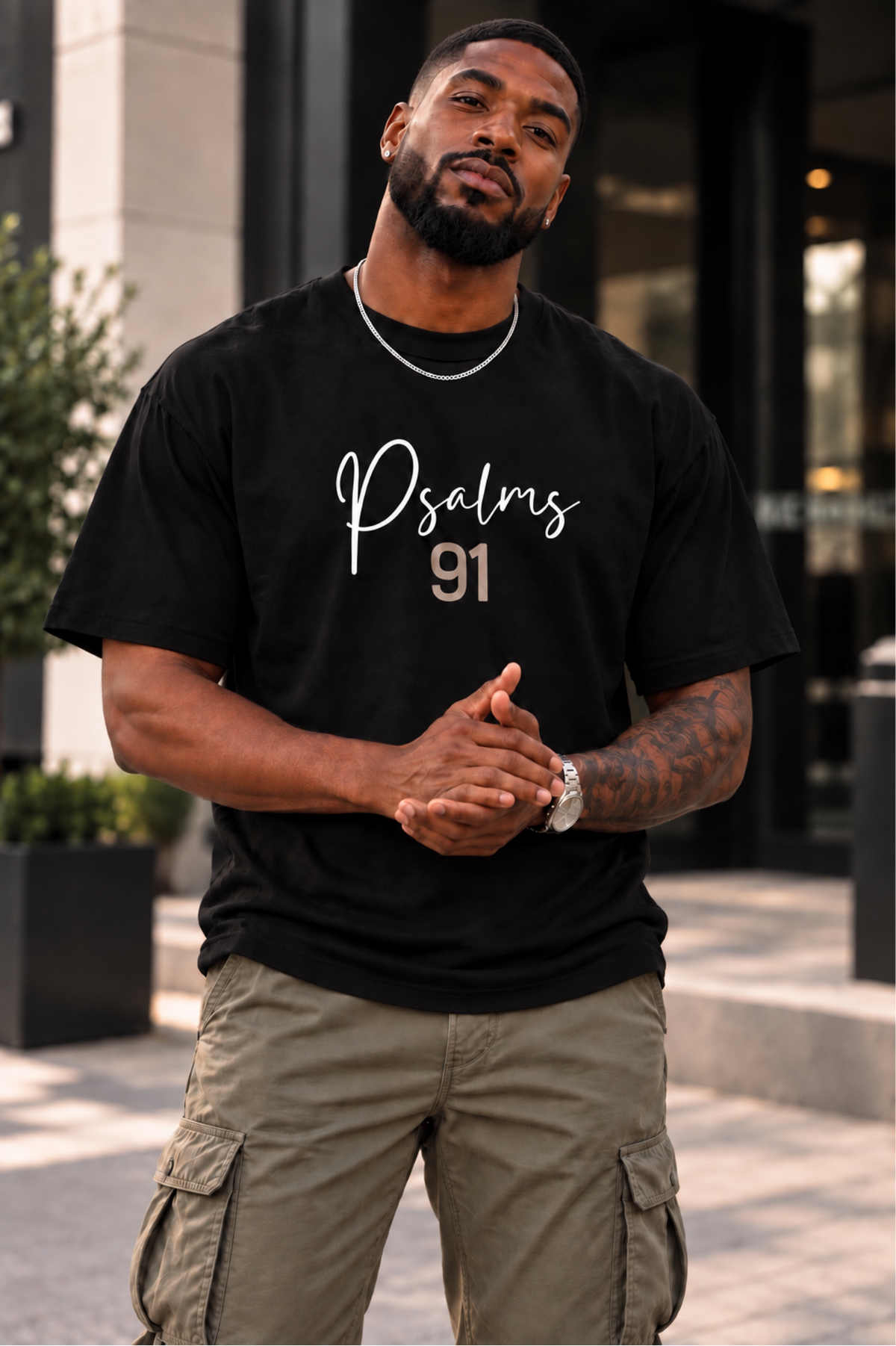 A man wearing a black graphic t-shirt with 'Psalms 91' printed on the front, paired with olive green cargo pants, standing outdoors in front of a modern building.