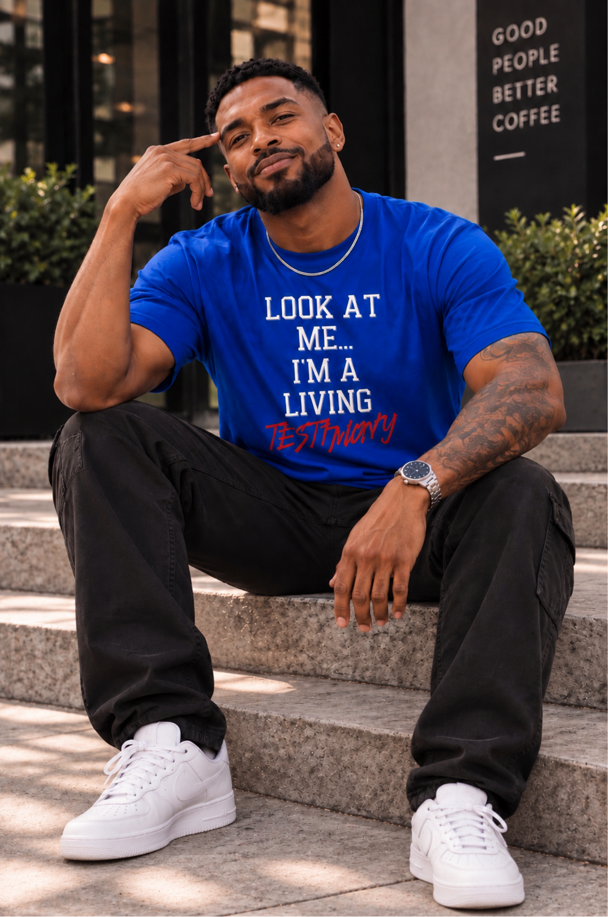 A man wearing a bright blue graphic t-shirt with white and red text, black cargo pants, and white sneakers, sitting on outdoor stone steps in front of a building.