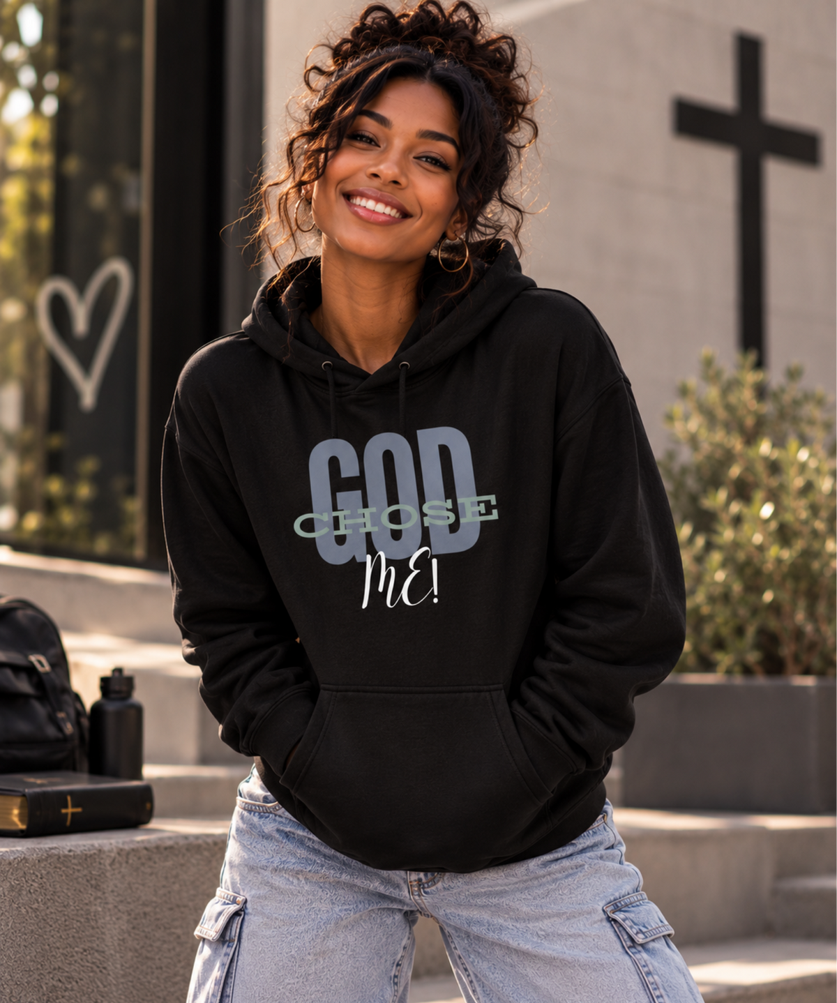A woman wearing a black pullover hoodie with the text 'GOD CHOSE ME' printed on the front, paired with light-wash cargo jeans, posing outdoors in front of a stone wall with a cross.