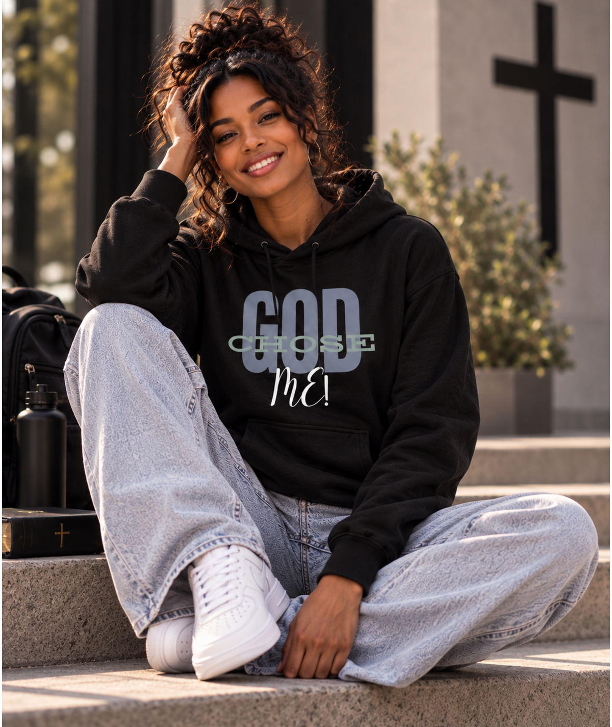 A woman sits on outdoor stone steps wearing a black hoodie with the text 'GOD CHOSE Me!' in gray and white, paired with light-wash wide-leg jeans and white sneakers.