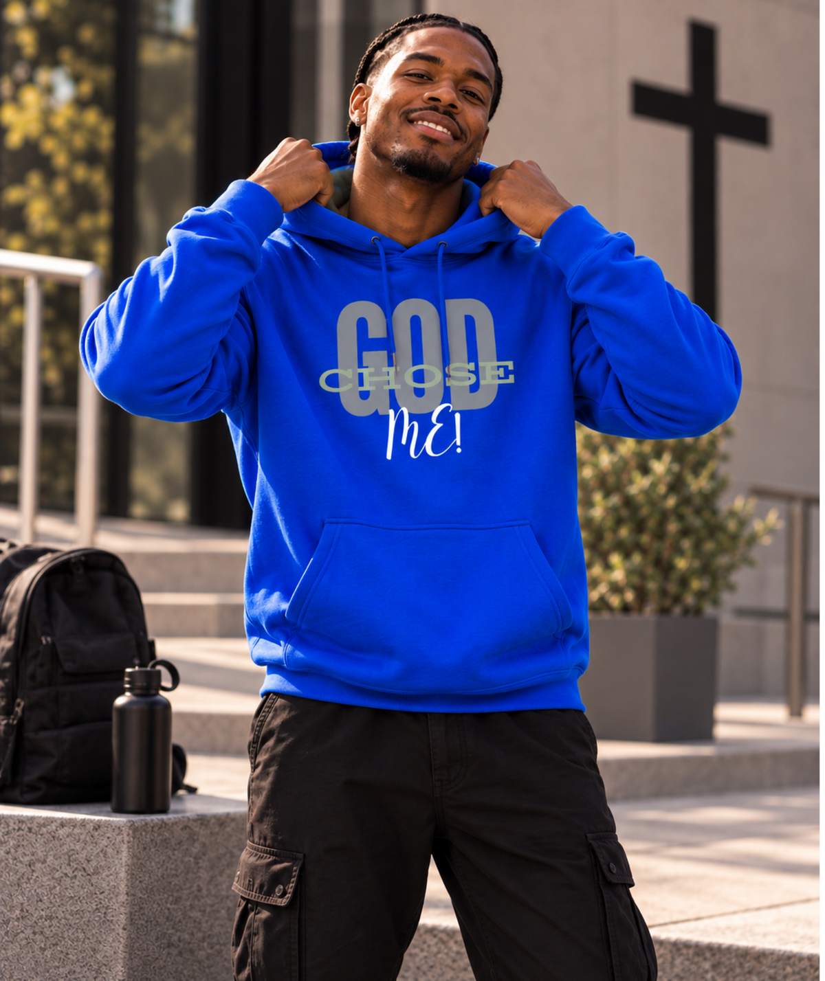 A man wearing a bright blue hoodie with 'GOD CHOSE ME!' printed on the front, standing outdoors in front of a church building. A black backpack and water bottle sit on a stone ledge beside him.