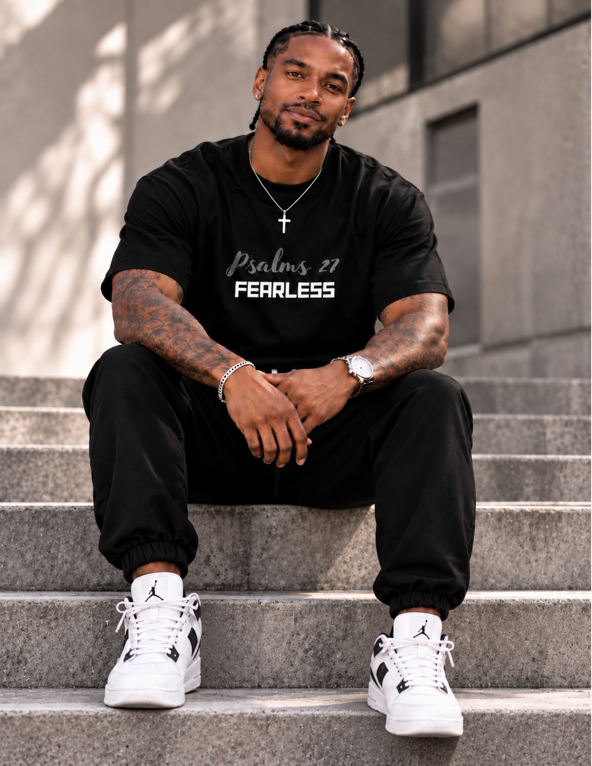 A man sits on concrete steps wearing a black t-shirt with 'Psalm 27 FEARLESS' printed on the front, black jogger pants, and white sneakers with black accents.