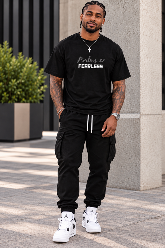 A man wearing a black crewneck t-shirt with 'Psalms 37 FEARLESS' printed in white on the chest, paired with black cargo jogger pants and white sneakers, standing outdoors in front of a modern stone column.