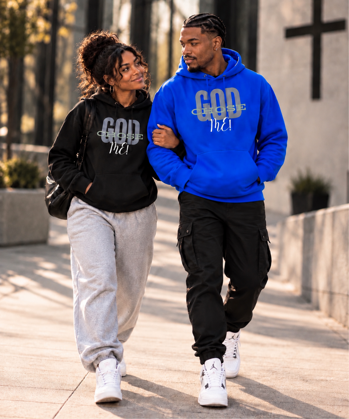 A couple walking arm-in-arm outdoors on a sunny day. The woman wears a black hoodie with 'GOD OVER ME' in white text, paired with light gray sweatpants and white sneakers.